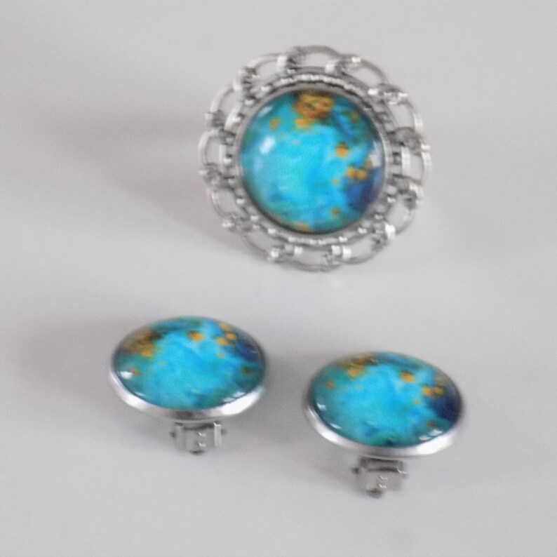 May include: A silver ring, a pair of earrings, and a brooch with a blue and gold design. The jewelry features a round, cabochon-shaped stone with a swirling blue and gold pattern.