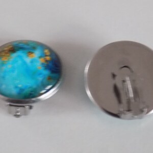 May include: A pair of silver clip-on earrings with a round blue and gold design. The earrings have a silver metal backing.