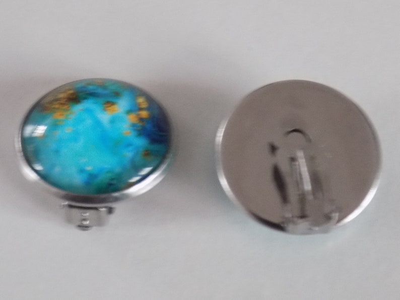 May include: A pair of silver clip-on earrings with a round blue and gold design. The earrings have a silver metal backing.