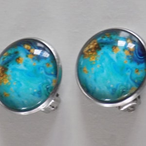 May include: A pair of silver clip-on earrings with blue and gold abstract designs. The earrings have a round shape and a smooth, shiny finish.