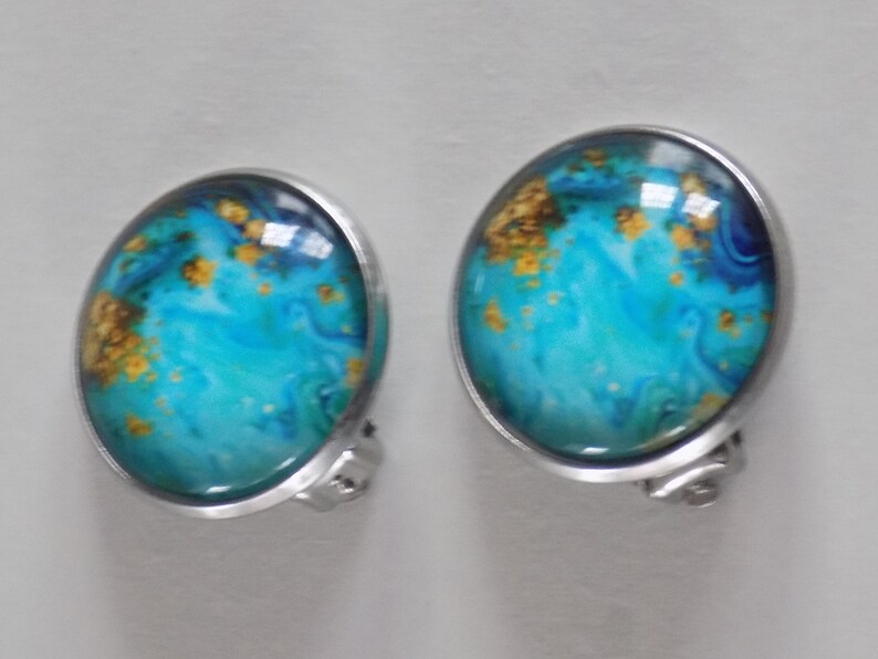 May include: A pair of silver clip-on earrings with blue and gold abstract designs. The earrings have a round shape and a smooth, shiny finish.