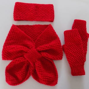 Op de afbeelding: Een felrode gebreide winterset, bestaande uit een hoofdband, een sjaal en vingerloze handschoenen. De set is gemaakt van een textuurgebreide stof. De sjaal heeft een uniek strikachtig ontwerp. De handschoenen zijn open bij de vingers.