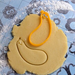 May include: A yellow plastic cookie cutter in the shape of an eggplant is on a blue and white patterned surface covered in white flour. The cutter is on top of a round piece of cookie dough.