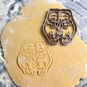 May include: A brown plastic cookie cutter in the shape of two hugging animals. The cutter is on top of a round piece of dough, ready to be used to make cookies.