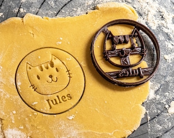 Puede incluir: Un cortador de galletas marrón en forma de gato con el nombre "Jules" inscrito en él. El cortador está encima de una masa de galletas amarilla sobre una superficie negra.