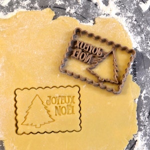 Peut inclure: Un emporte-pièce en plastique brun avec les mots "Joyeux Noël" et un motif de sapin de Noël. L'emporte-pièce est posé sur une feuille de pâte à biscuits jaune.