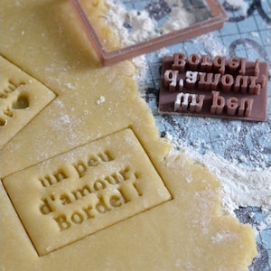 Könnte beinhalten: Nahaufnahme eines braunen Keksteigs mit einem rechteckigen Ausstecher, der in den Teig gedrückt wurde. Der Ausstecher trägt die Aufschrift "un peu d'amour. Bordel!"  Im Hintergrund ist ein Satz brauner Alphabet-Ausstecher zu sehen.