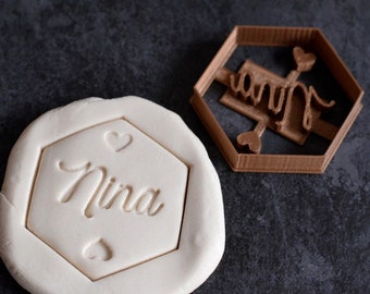 May include: A brown plastic cookie cutter in the shape of a hexagon with the word "Nina" and two hearts inside. The cutter is on a gray surface next to a white piece of clay with the cutter's impression.