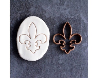 May include: A brown metal cookie cutter in the shape of a fleur-de-lis. The cutter is shown next to a piece of white modeling clay with the fleur-de-lis design pressed into it.