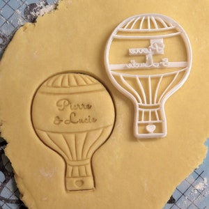 Puede incluir: Cortador de galletas de plástico blanco en forma de globo aerostático con el texto "Pierre & Lucie" en su interior. El cortador está sobre una masa de galletas de color marrón claro.