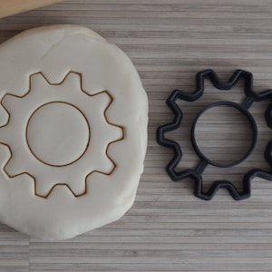 Puede incluir: Un cortador de galletas de plástico negro con forma de engranaje, junto a una masa de galletas blanca con un diseño de engranaje impreso en ella.