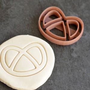 Puede incluir: Un cortador de galletas en forma de pretzel marrón junto a un trozo de masa con una impresión de pretzel. El cortador es de plástico y la masa es de color claro. La imagen está sobre una superficie gris oscuro.
