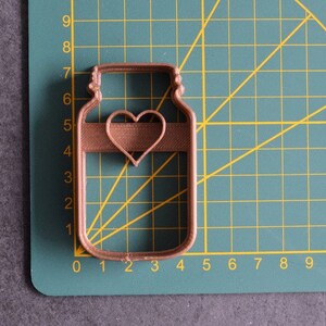 May include: A brown metal cookie cutter shaped like a mason jar with a heart cutout in the center.