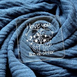 Puede incluir: Un posavasos de acrílico transparente con el texto "i love you like no otter" y una imagen de dibujos animados de dos nutrias sosteniendo corazones.