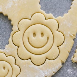 May include: A light beige rolled-out cookie dough sheet featuring two pressed flower-shaped impressions. Each flower shape has a central circular face with two small oval eyes and a curved mouth, creating a smiling expression in the dough.