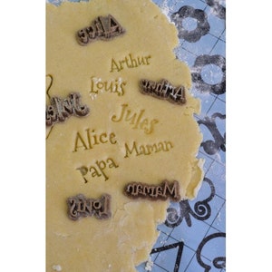 May include: Close-up of a cookie dough sheet with various names stamped into the dough, including "Arthur", "Louis", "Alice", "Papa", "Maman", "Jules", and "Mama".