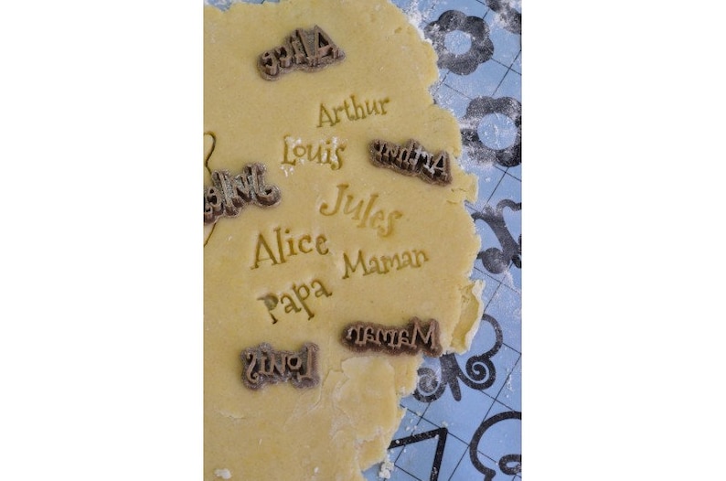 May include: Close-up of a cookie dough sheet with various names stamped into the dough, including "Arthur", "Louis", "Alice", "Papa", "Maman", "Jules", and "Mama".