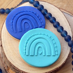 May include: Two blue and turquoise rainbow-shaped silicone stamps with a small heart in the center of each rainbow. The stamps are on a wooden surface.