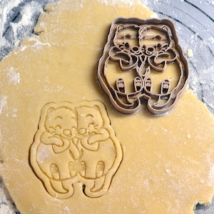 May include: Two brown cookie cutters, one of two otters holding each other and the other of two otters facing each other, on a light brown surface covered in white flour.