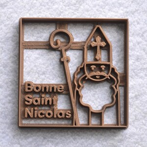 May include: A brown plastic cookie cutter in a square shape. The design features a depiction of Saint Nicholas with a staff and a hat with a cross. The words "Bonne Saint Nicolas" are printed on the left side of the cutter.