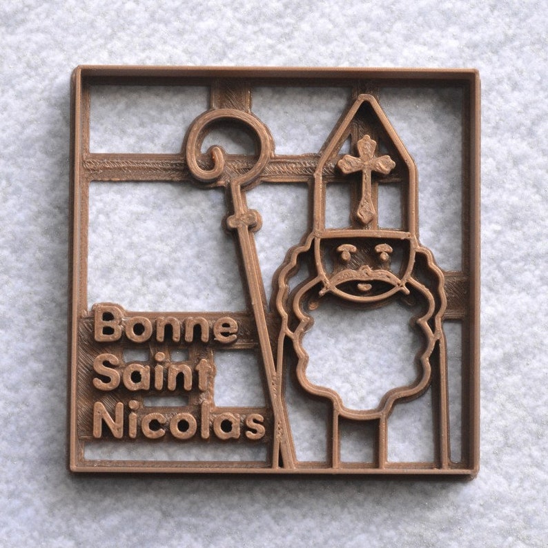 May include: A brown plastic cookie cutter in a square shape. The design features a depiction of Saint Nicholas with a staff and a hat with a cross. The words "Bonne Saint Nicolas" are printed on the left side of the cutter.