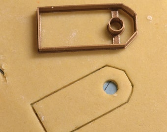 May include: A brown plastic cookie cutter shaped like a tag with a hole in the center. The cutter is on a light brown surface.