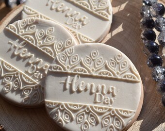 May include: Three white round cookies with a decorative design and the text "Henna Day" on each. The cookies are on a wooden surface.