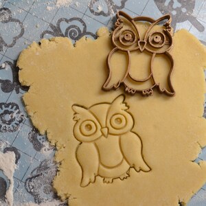 May include: Two brown metal owl cookie cutters on a light blue surface covered in white flour. One cutter is on top of a round piece of cookie dough, the other is next to it.