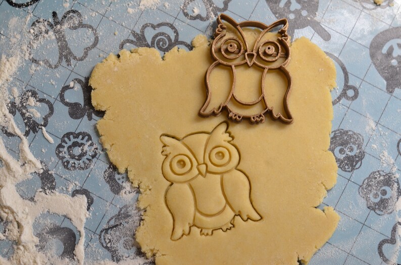 May include: Two brown metal owl cookie cutters on a light blue surface covered in white flour. One cutter is on top of a round piece of cookie dough, the other is next to it.