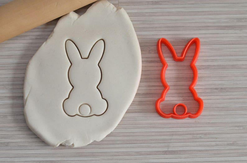 Puede incluir: Un cortador de galletas de pl&aacute;stico rojo con forma de conejo. El cortador est&aacute; sobre una superficie blanca con un rodillo y una masa.