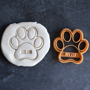 May include: A brown plastic cookie cutter in the shape of a paw print with the name "Kelvin" inscribed inside. The cutter is next to a white fondant cookie with the same paw print design.