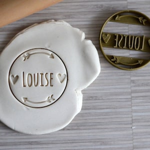 May include: A gold-colored cookie cutter with the name "LOUISE" in a circle with arrows and hearts. The cutter is on a white surface with a rolling pin in the background.
