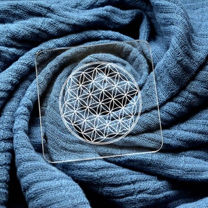 May include: A white geometric flower of life design on a clear acrylic coaster. The coaster is on a blue fabric background.