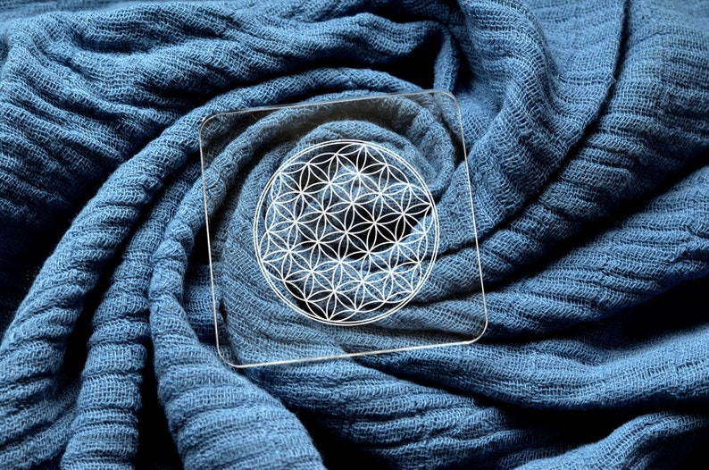 May include: A white geometric flower of life design on a clear acrylic coaster. The coaster is on a blue fabric background.