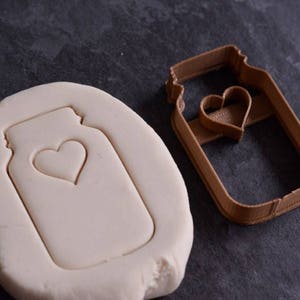 May include: A brown plastic cookie cutter in the shape of a mason jar with a heart cutout. A white clay cookie is shown with the cookie cutter imprint.