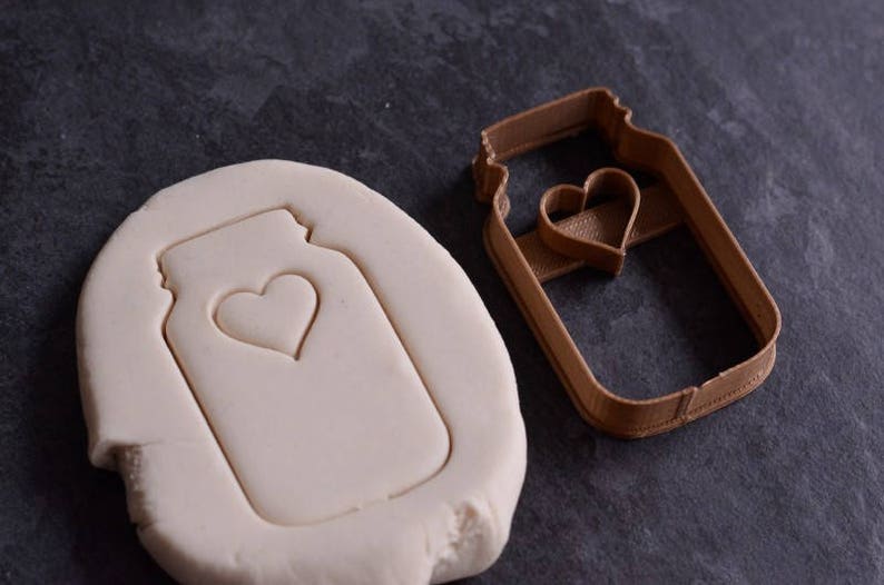 May include: A brown plastic cookie cutter in the shape of a mason jar with a heart cutout. A white clay cookie is shown with the cookie cutter imprint.