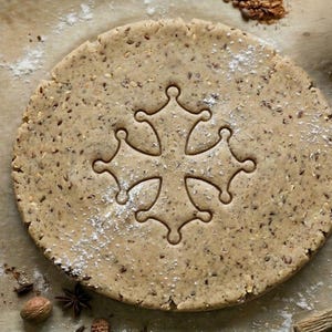 Peut inclure: Une pièce ronde et plate de pâte à biscuit beige avec une texture granuleuse visible et des graines sombres éparpillées. Une croix occitane est embossée au centre de la pâte. La surface est légèrement saupoudrée de farine blanche.