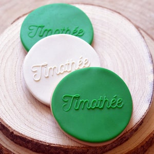 May include: Three round cookies, two green and one white, with the name "Timothee" written in white icing on the green cookies and in green icing on the white cookie.