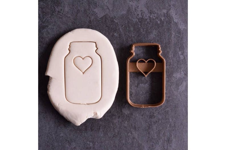 May include: A brown plastic cookie cutter in the shape of a mason jar with a heart cutout. The cookie cutter is on a gray surface next to a white cookie dough imprint of the same shape.