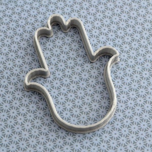 Può includere: Un tagliabiscotti color argento a forma di mano con un pollice e quattro dita. Il tagliabiscotti è in metallo e ha una superficie strutturata. Lo sfondo è un motivo azzurro.