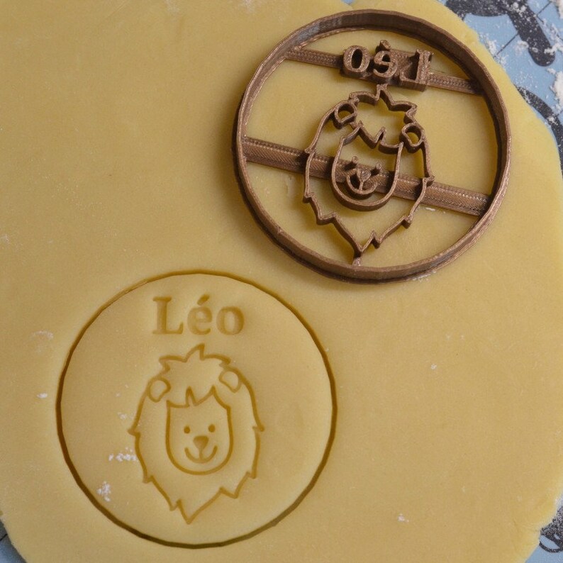 Pode incluir: Um cortador de biscoitos de pl&aacute;stico marrom com um rosto de le&atilde;o e o nome "Leo" escrito nele. O cortador est&aacute; em cima de uma massa com o mesmo desenho impresso nela.