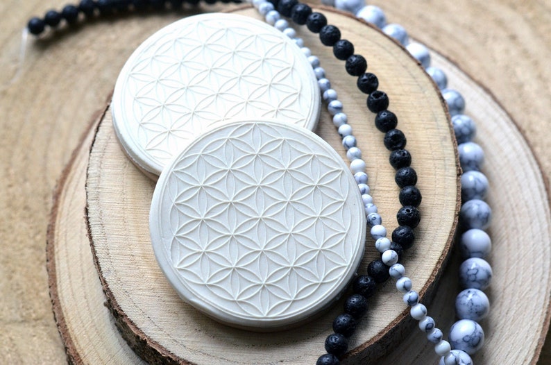May include: Two white round coasters with a raised geometric flower of life design. The coasters are on top of two wooden slices. A black lava rock beaded necklace and a white and gray beaded necklace are on top of the wooden slices.