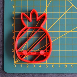 May include: A red plastic pineapple-shaped cookie cutter with a smiling face and diagonal lines. The cutter is on a green cutting mat with a grid and measurement markings in inches.