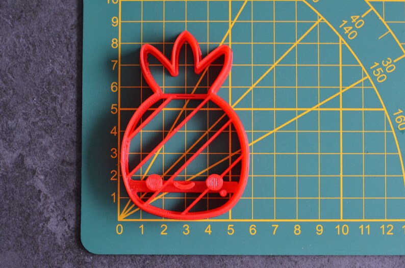 May include: A red plastic pineapple-shaped cookie cutter with a smiling face and diagonal lines. The cutter is on a green cutting mat with a grid and measurement markings in inches.