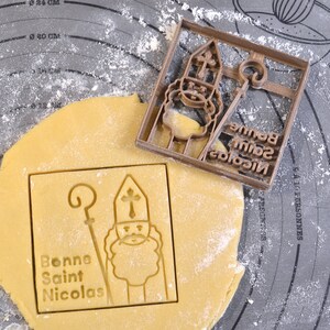 May include: A brown plastic cookie cutter depicting Saint Nicholas, with the text "Bonne Saint Nicolas". The cutter sits on a sheet of dough, with an imprint of the saint and text. The dough is on a grey surface.