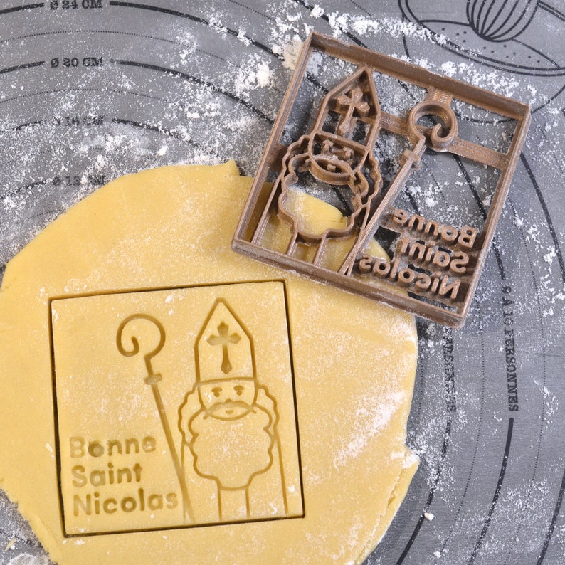 May include: A brown plastic cookie cutter depicting Saint Nicholas, with the text "Bonne Saint Nicolas". The cutter sits on a sheet of dough, with an imprint of the saint and text. The dough is on a grey surface.