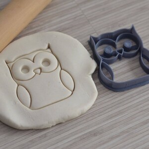 May include: A gray plastic owl cookie cutter with a white dough imprint of the owl. The cookie cutter is on a wooden surface.