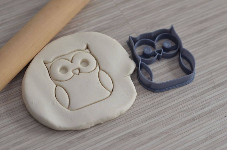 May include: A gray plastic owl cookie cutter with a white dough imprint of the owl. The cookie cutter is on a wooden surface.