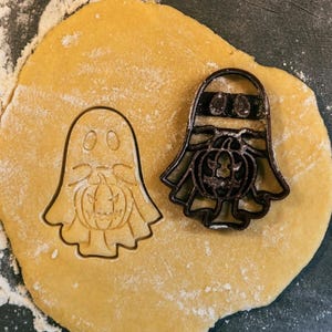May include: A close-up of a cookie cutter set for Halloween. The cutters are shaped like a ghost and a pumpkin. The cutters are on a surface covered in flour.