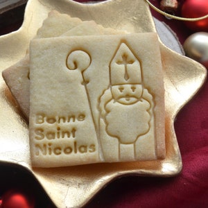 May include: Square-shaped shortbread cookies with an embossed image of Saint Nicholas holding a staff, and the words "Bonne Saint Nicolas". The cookies are light brown and rest on a gold star-shaped plate.